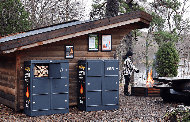 Piffl Brasved – Hållbar ved för dina skogsäventyr Vindskydd i skogen med två Piffl Brasved boxar och en tjej som värmer sig vid elden.