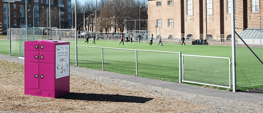 Spela fotboll i Norrköping – Låna en boll från Pifflboxen Piffl aktivitetsbox vid en fotbollsplan i Norrköping där barn spelar och har roligt