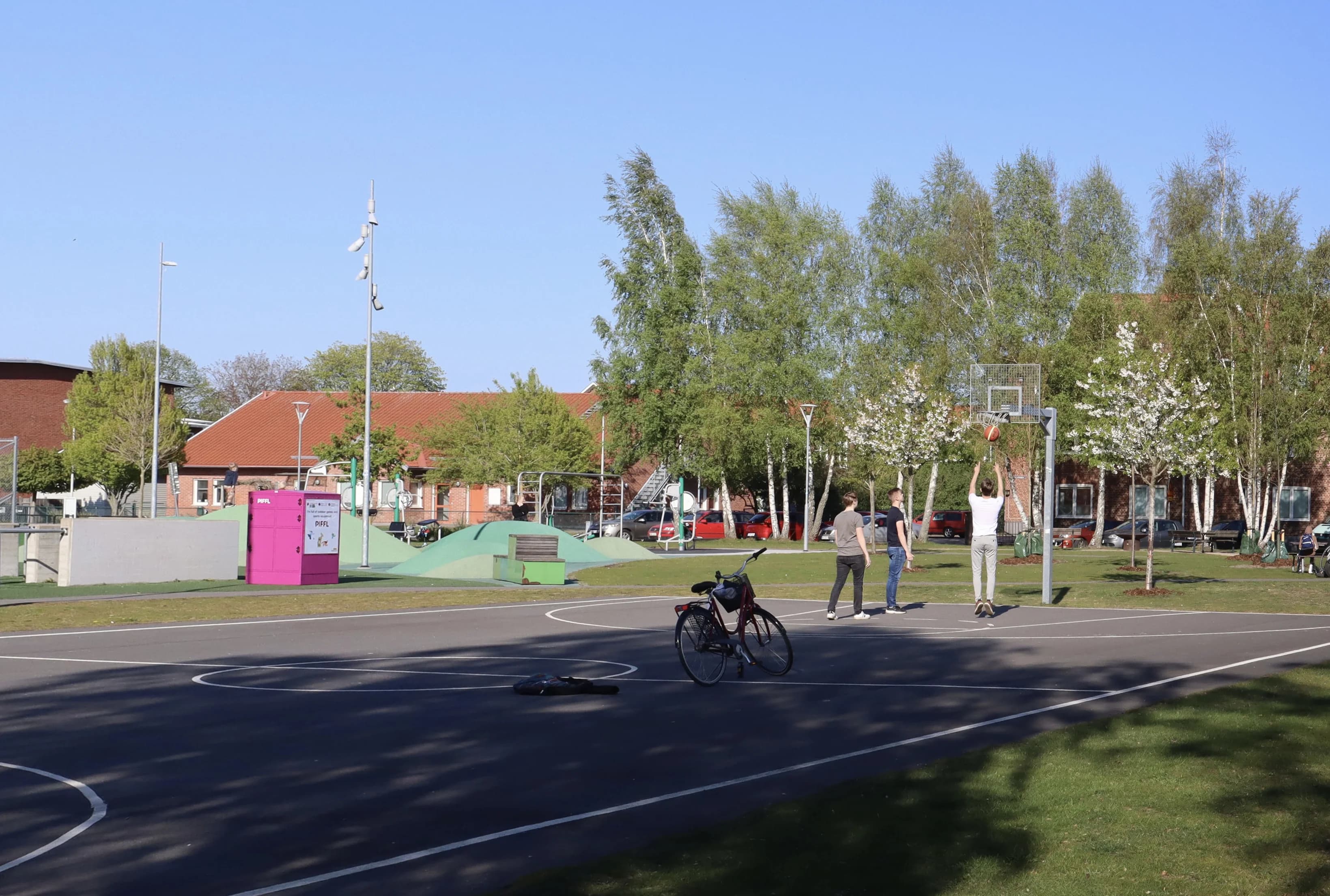 En rosa Pifflbox vid en utomhusplan där tre personer spelar basket